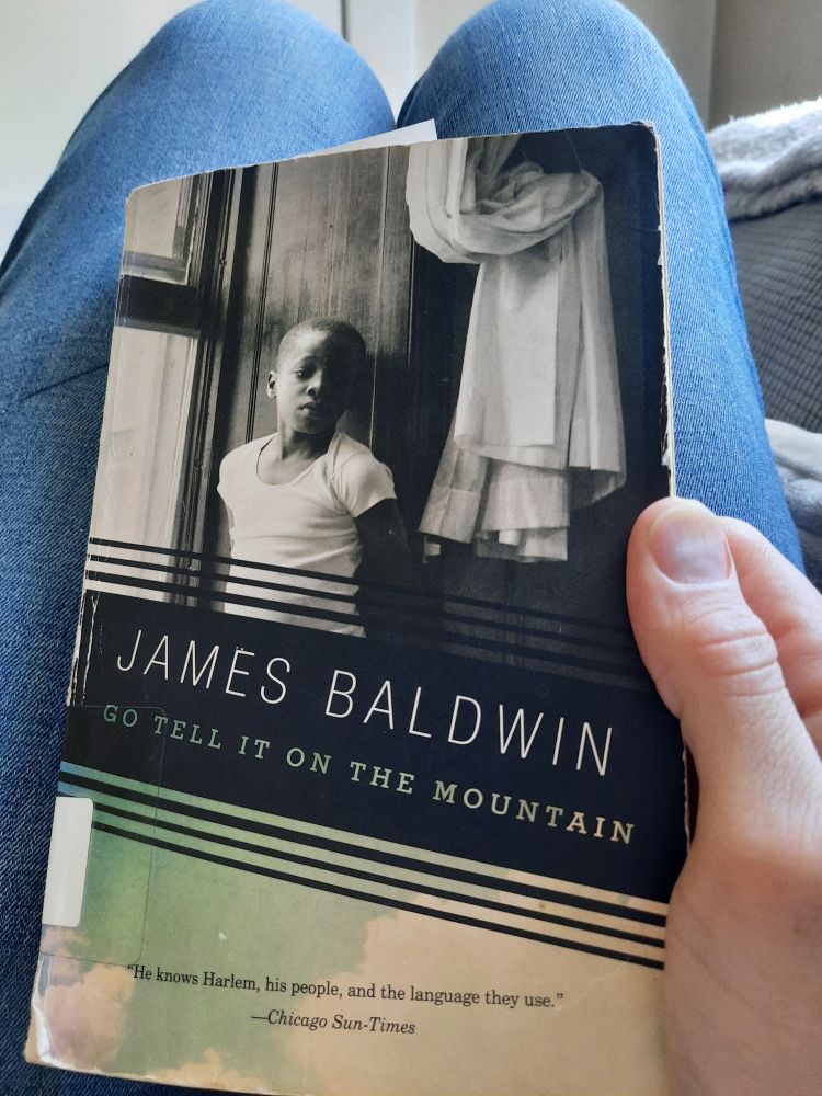 A copy of GO TELL IT ON THE MOUNTAIN by James Baldwin. The cover photograph is  boy, copyright Leonard Freed/Magnum Photos. It depicts a young Black boy standing against a wall between a window and curtain. He has a sad look in his eyes, and his face is half-covered in shadow.
