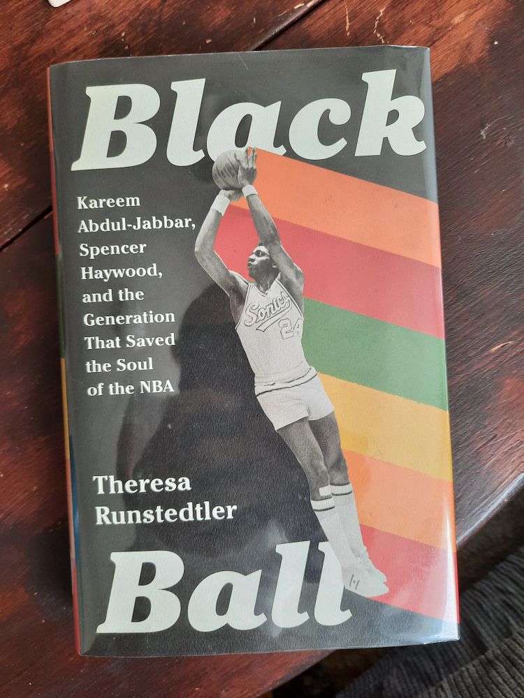 Text: BLACK BALL: KAREEM ABDUL-JABBAR, SPENCER HAYWOOD, AND THE GENERATION THAT SAVED THE SOUL OF THE NBA by Theresa Runstedtler 

Image: a black and white photo of Spencer Haywood, wearing a #24 Sonics jersey, holding a basketball above his head. Half the book cover is black, and behind Haywood are stripes representing the Pan-African flag 