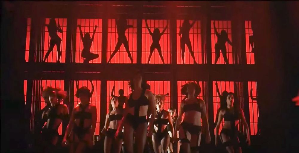 Cell block tango from the movie musical Chicago - a musical number showing women’s silhouettes against a red backdrop behind bars, women in the ground level dance in black bodysuits and dance wear 