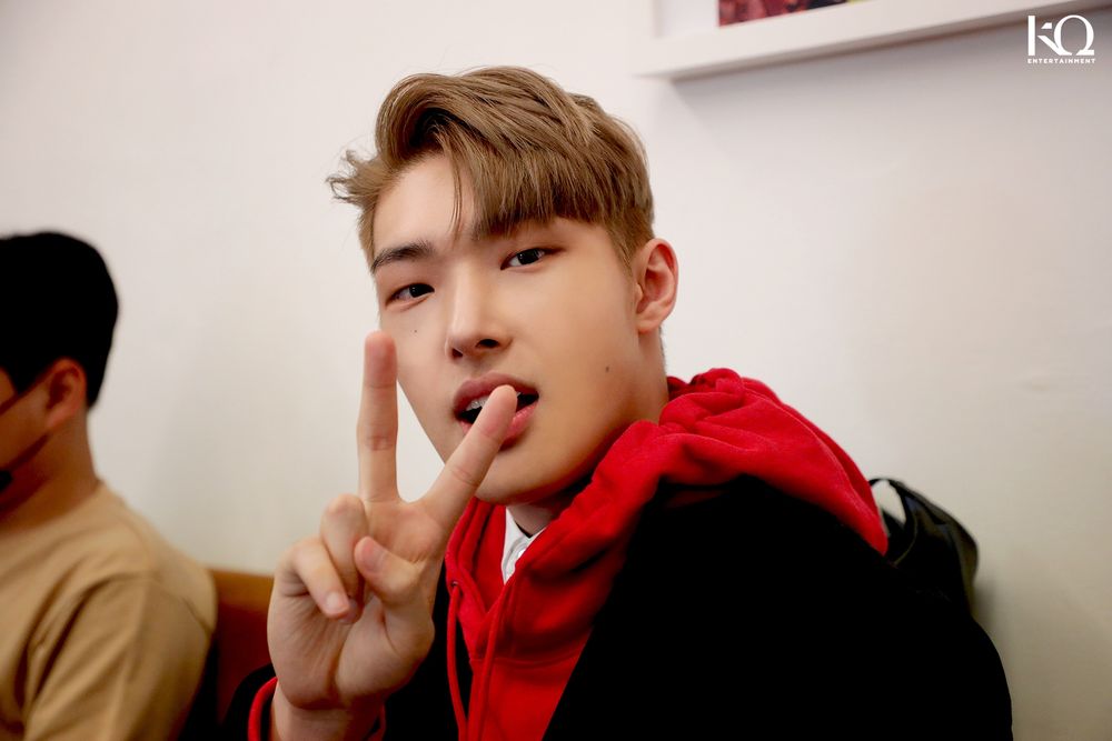 Song Mingi (ATEEZ) in a red hoodie and jacket making a peace sign at the camera