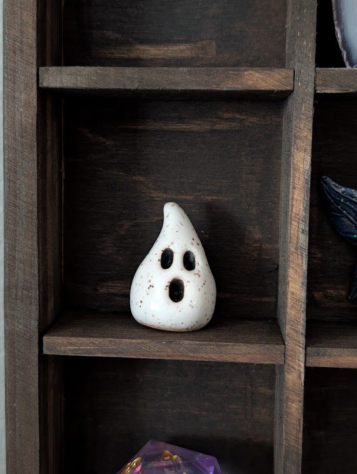 a photo of a tiny ceramic ghost, about 1 inch tall. teardrop shaped, painted speckled white, with three black ovals for eyes and mouth