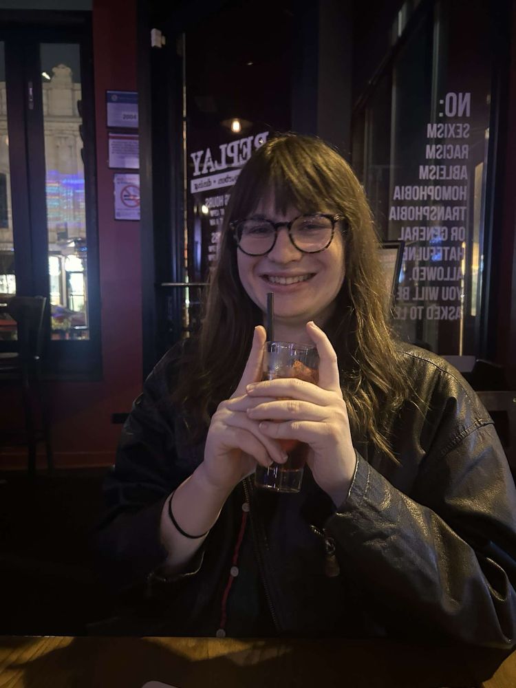 Beatrice in a bar holding a drink with both hands and smiling