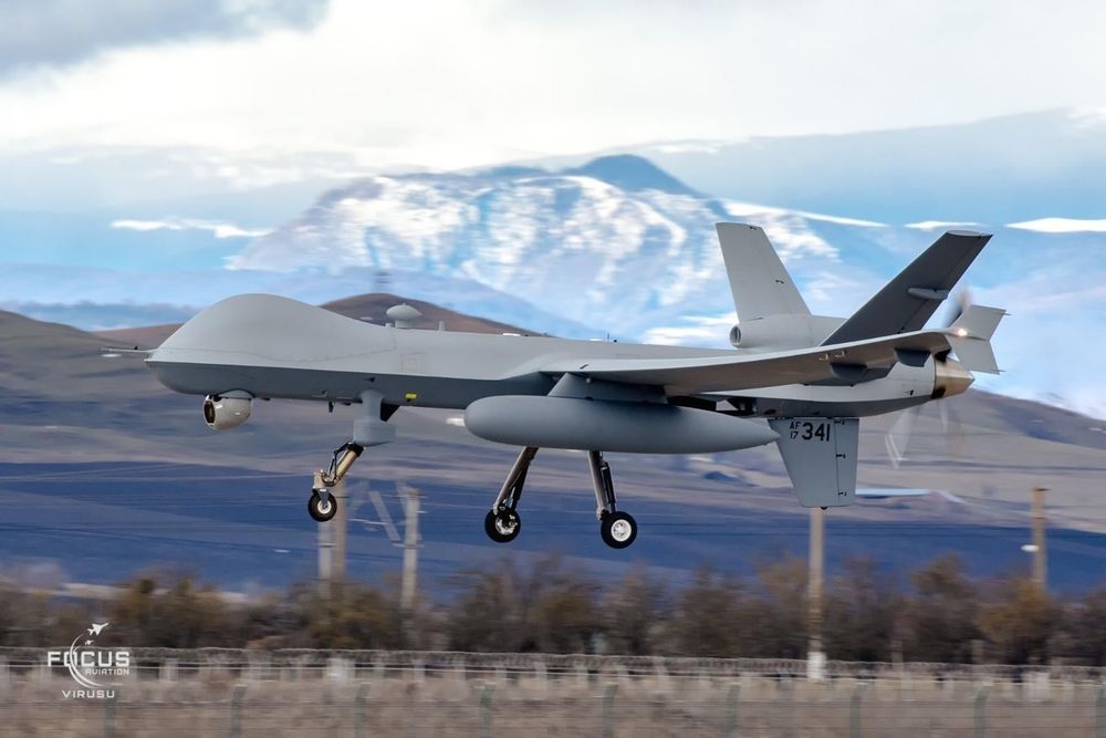 US Air Force MQ-9A 17-4341 on approach to land at Campia Turzii, RO. Picture shows an uncrewed aircraft against a blurry background of hills and mountains on a partly cloudy day. Text on the tail of the aircraft says "AF 17 341" in black letters.