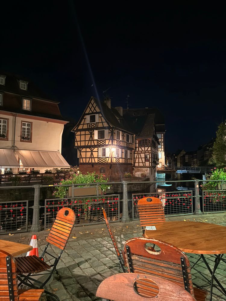 Utsikt mot et gult, eventyraktig hus langs vannet. Tomme bord og stoler i forkant, og et gjerde som skiller uteplassen fra elva. Gjerdet er fylt med røde, hjerteformede hengelåser. Himmelen er mørk. 