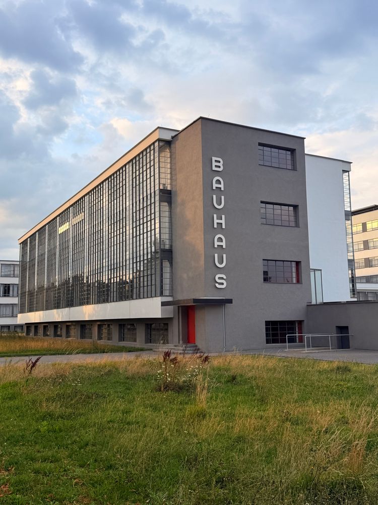 Modernistisk bygning i hvit og grå murpuss, med en stor tre-etasjers glassfasade til venstre og teksten ‘BAUHAUS’ stående vertikalt på den mørkegrå veggen nærest oss i bildet. Solnedgang, med lett skydekke. Grønt gress og høye gule strå i forgrunn. 