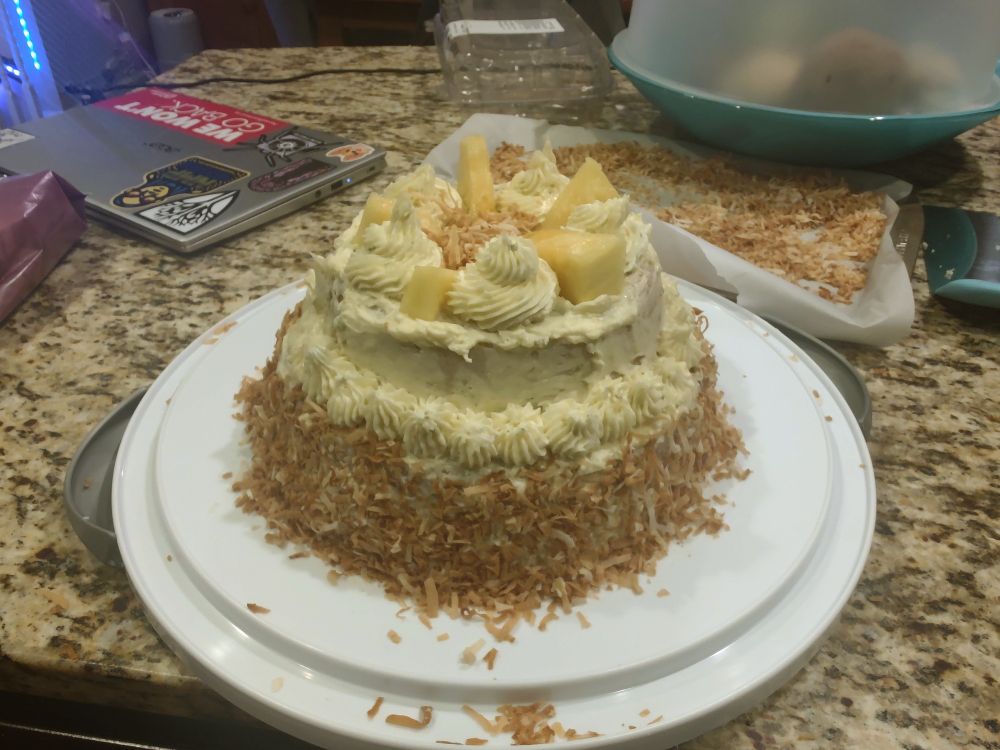 A two tiered cake with yellow frosting.  There is toasted coconut on the bottom layer and pineapple on the top layer