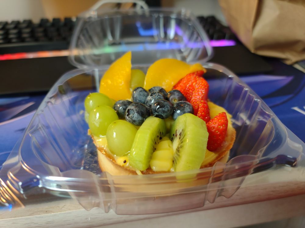 A miniature fruit tart covered with kiwi, strawberries, blueberries, melon, and grapes.  It is sitting in a small plastic carrier.