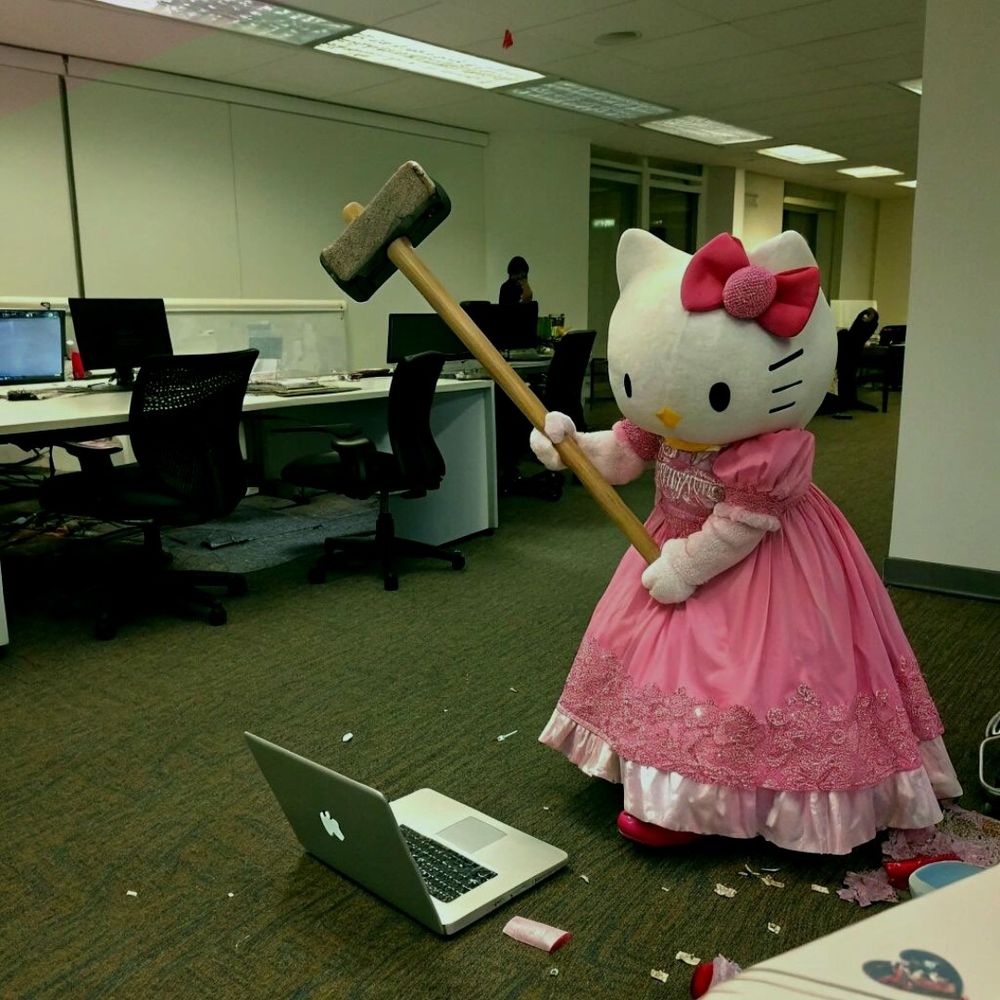 Someone dressed as Hello Kitty in a pink princess dress wielding a sledgehammer standing over a laptop on the ground. 