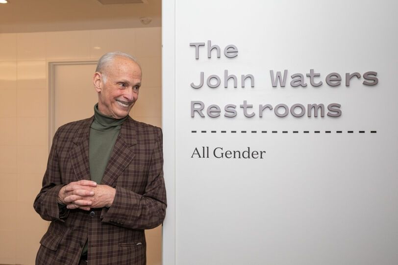 John Waters dressed in a brown plaid sport coat and gray turtleneck leaning against a white wall that says in gray letters "The John Waters Restrooms all gender".