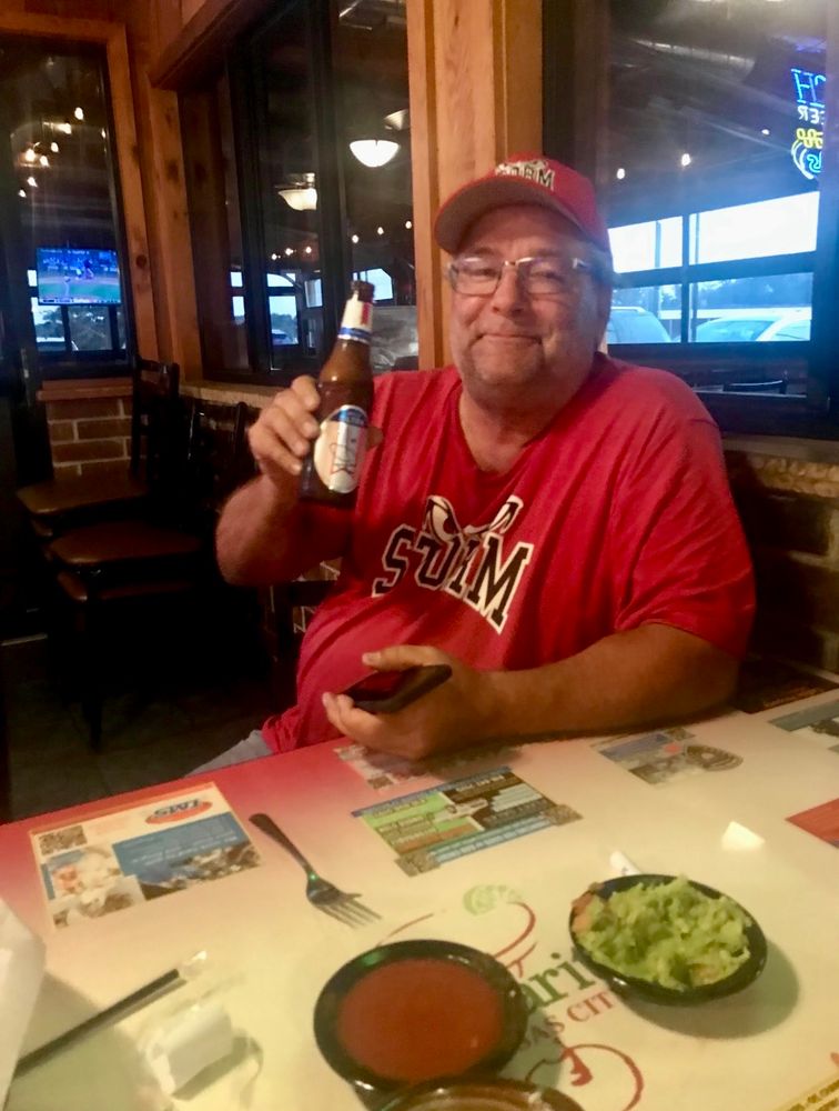 At the dinner table at Margaritas restaurant, Dad wears his Storm baseball gear (hat and shirt) in red and toasts a michelob ultra (drank under protest when he had to cut back on carbs)
