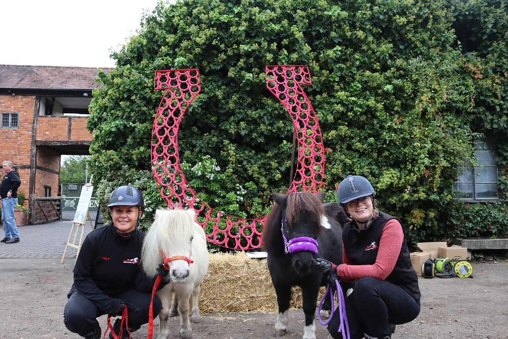 Redwings Shetlands Tinkerbell and Marjorie in front of a giant pink horseshoe with their handlers Hollie and Jude.