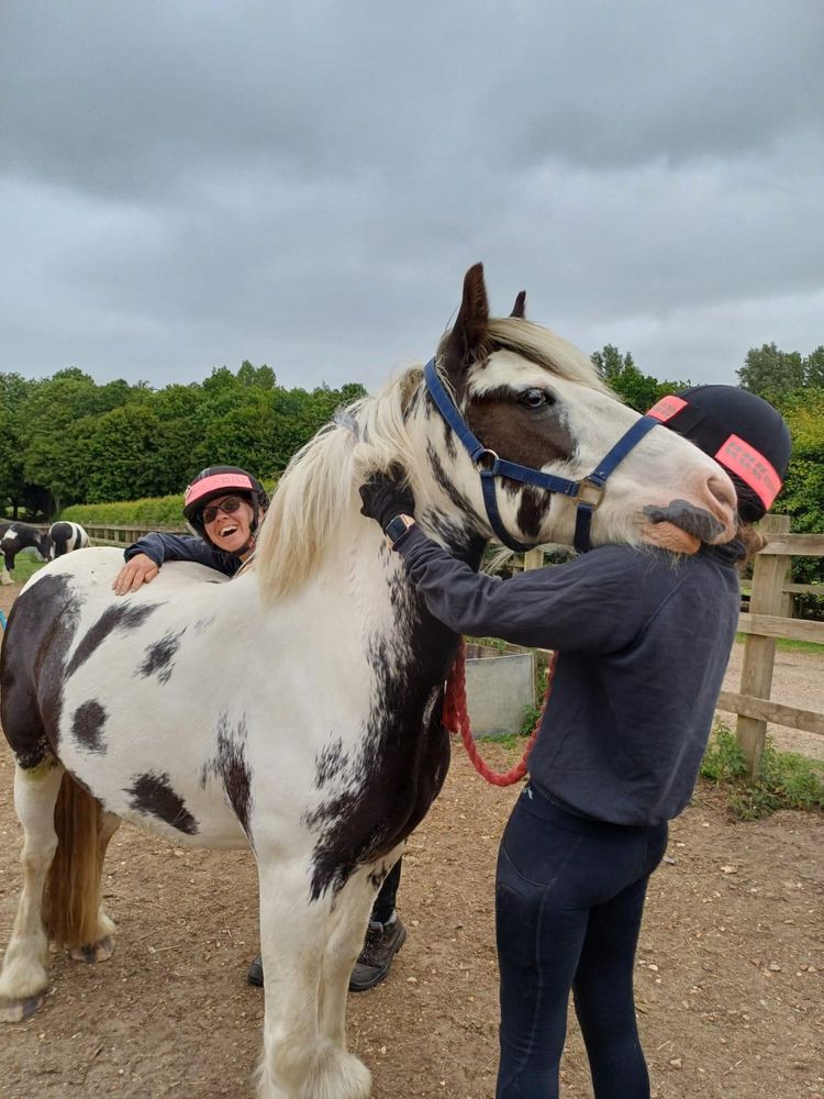 A black and white pony is enjoying a hug and scratch from one of his carers whilst another carer smiles at the camera over the back of the pony.
