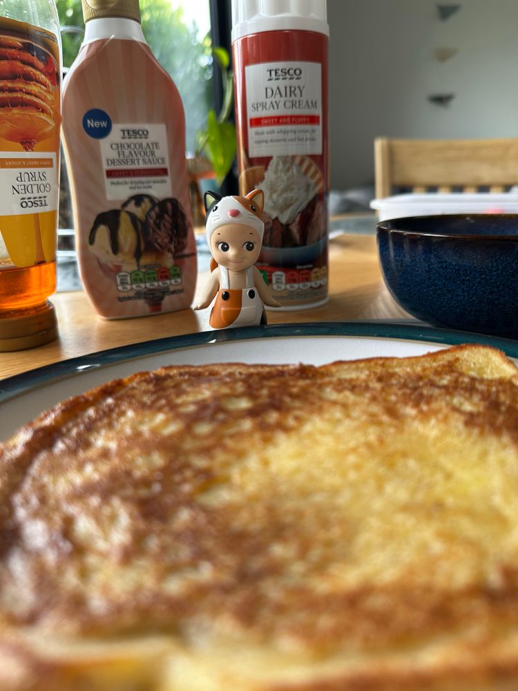 A calico Sonny Angel figure (small doll with cat ears) is standing in front bottles of golden syrup and chocolate sauce and a can of squirty cream. The Sonny Angel is looking at a piece of French toast. The angle makes the toast look huge. 
