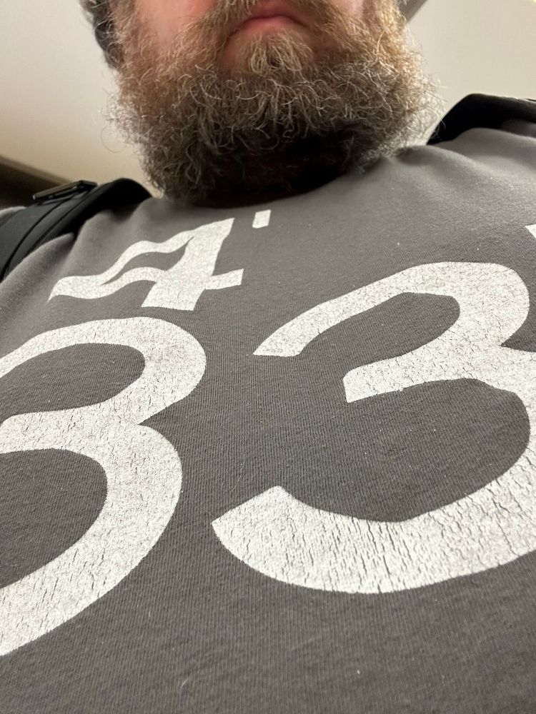 A close-up photo of a bearded man, wearing a dark gray t-shirt with a reference to John Cage's seminal avant-garde musical piece, 4′33”.