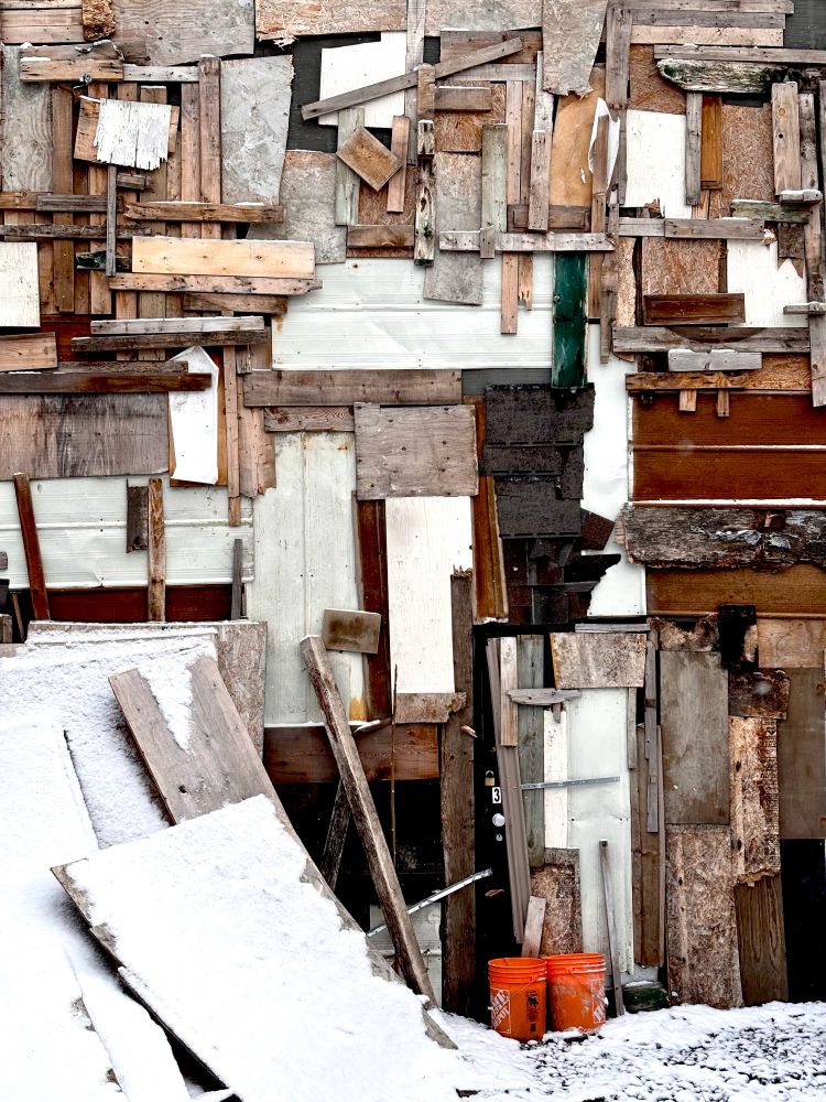 Photo of a building’s exterior wall absolutely covered in a patchwork of small scraps of lumber and plywood, with two bright orange construction buckets sitting on the snowy ground in front, contrasting against all the shades of brown and grey behind
