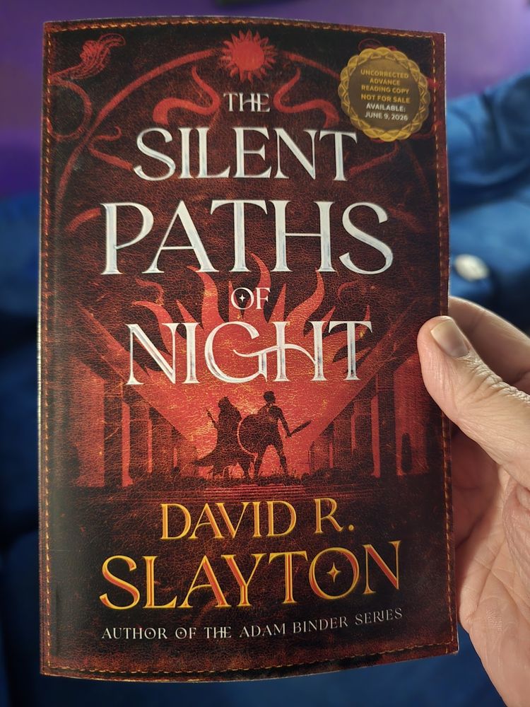 David holding an advanced reader copy (ARC) of The Silent Paths of Night, with a red cover and silhouettes of two figures, one a knight with a shield and a rogue in a cloak with a knife.