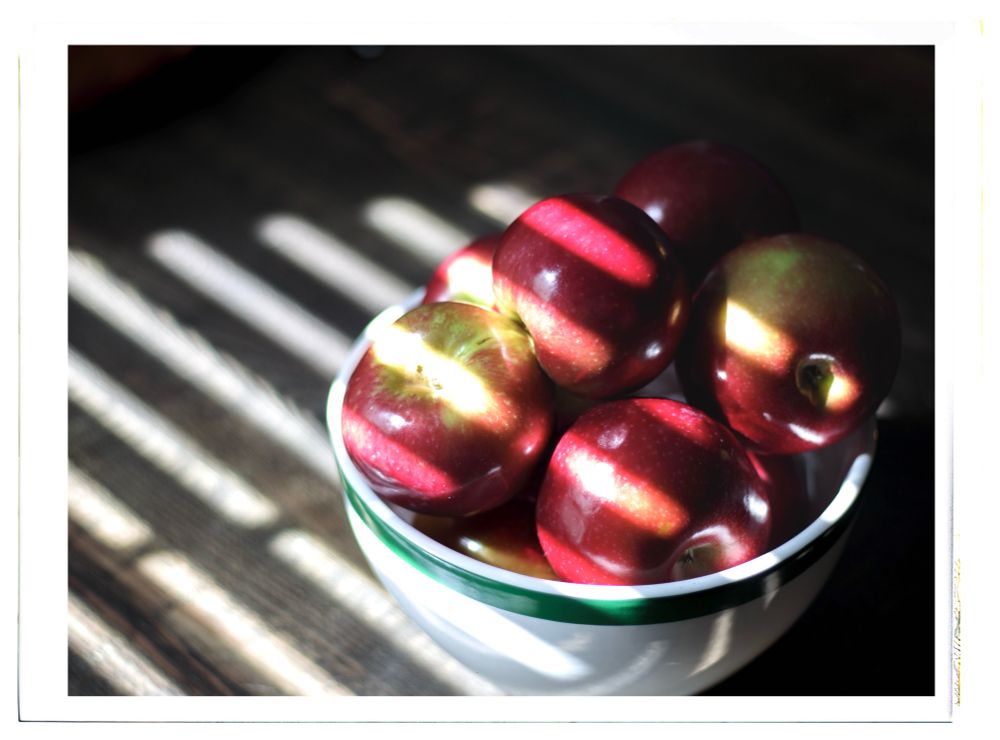 A bowl of red apples on a wood table with light from blinds shining through 