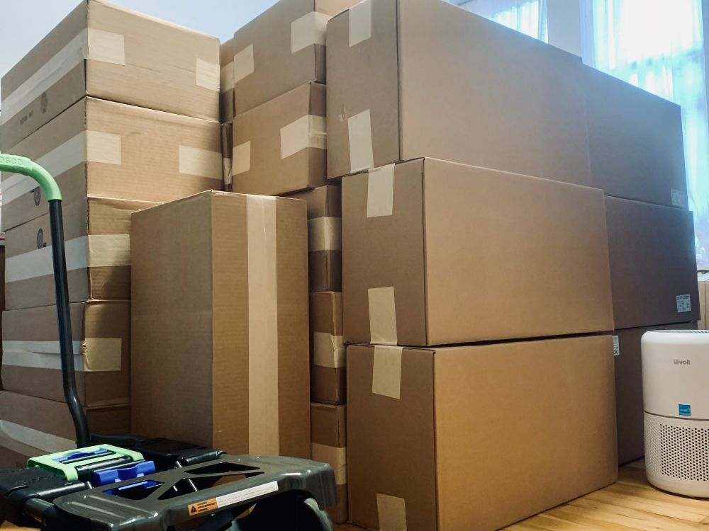 Stacks of cardboard boxes filled with rapid antigen tests for covid 19 acquired from the government sit in a living room on hardwood floors in front of open windows with white curtains. A white air purifier is on the ground to the right of the boxes and a grey metal rolling cart with a green handle is to the left.