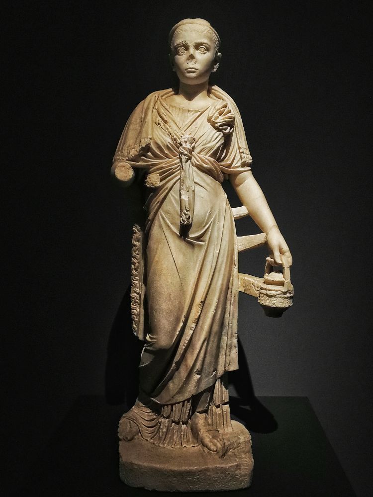 STATUE OF A PRIESTESS OF ISIS, C. 200 CE. PALAZZO CIAMPOLI, TAORMINA

Ancient Tauromenium was the centre of a major cult centre of the Roman Egyptian gods Isis and Serapis, one of the most important centres in the Mediterranean. None of the Serapeum survives today, but this statue and various other pieces of decoration have been found in excavations near the church of S. Pancrazio. This statue has a particular delicacy and grace in its carving. The priestess of Isis is identifiable by the famous Isaic knot in her dress, between her breasts. Her broken right arm would have held a sistrum or ceremonial rattle, and her left hand holds a rounded cista which contained items and substances used in the initiation of new members into the mystery cult of Isis. 