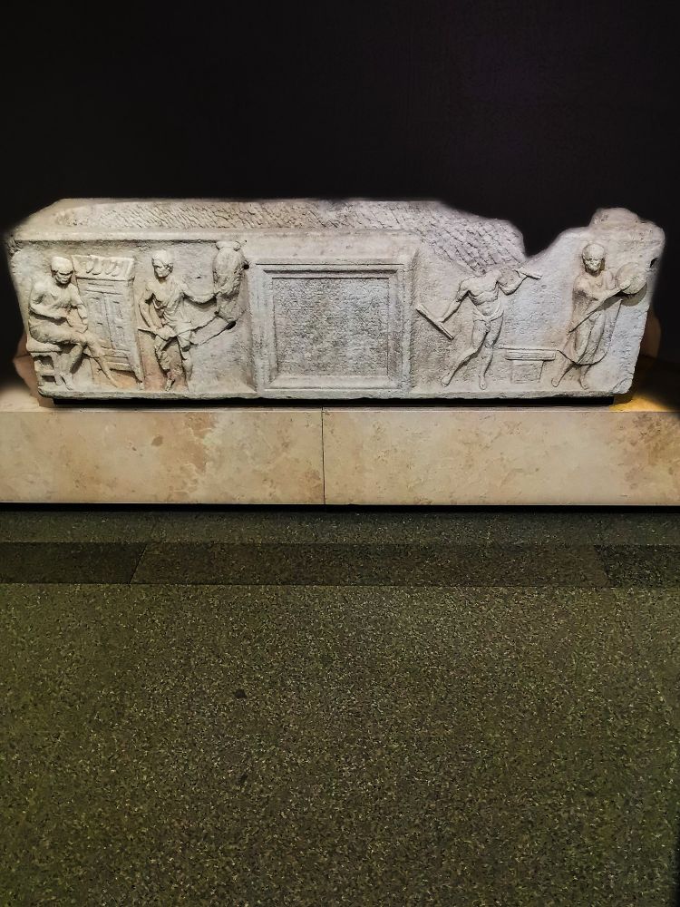 SARCOPHAGUS OF TITUS FLAVIUS TROPHIMAS, C. 1OO-140 CE. BATHS OF DIOCLETIAN

This interesting sarcophagus shows that artisans could have bespoke sarcophagi, which were not exclusively for the rich. At centre is a tabula with an inscription in Greek explaining that this is the coffin of T. Flavius Trophimas, a Greek Roman citizen (perhaps an imperial freedman) from Ephesus, "panmousas" or a cultivator of every art. It was commissioned by his friends Lucius Atilius Artemas and Claudia Apphias, both also of clear Greek origin. To the right of the tabula is a scene with two musicians, the one at far right beating a tambourine, the other in only a loincloth, playing a double flute and holding another flute. Both musicians are dancing in time with each other. On the other side of the tabula, a man is winding a rope from hanging skeins of fibre, while at far left is the deceased, seated as he makes a shoe, with a cupboard topped with wooden shoe-forms suggests his workshop. The inclusion of music in this sarcophagus relief suggests that the deceased, and/or his friends, were devotees of Isis, as the instruments being played frequently form part of representations of Isaic ceremonies. 