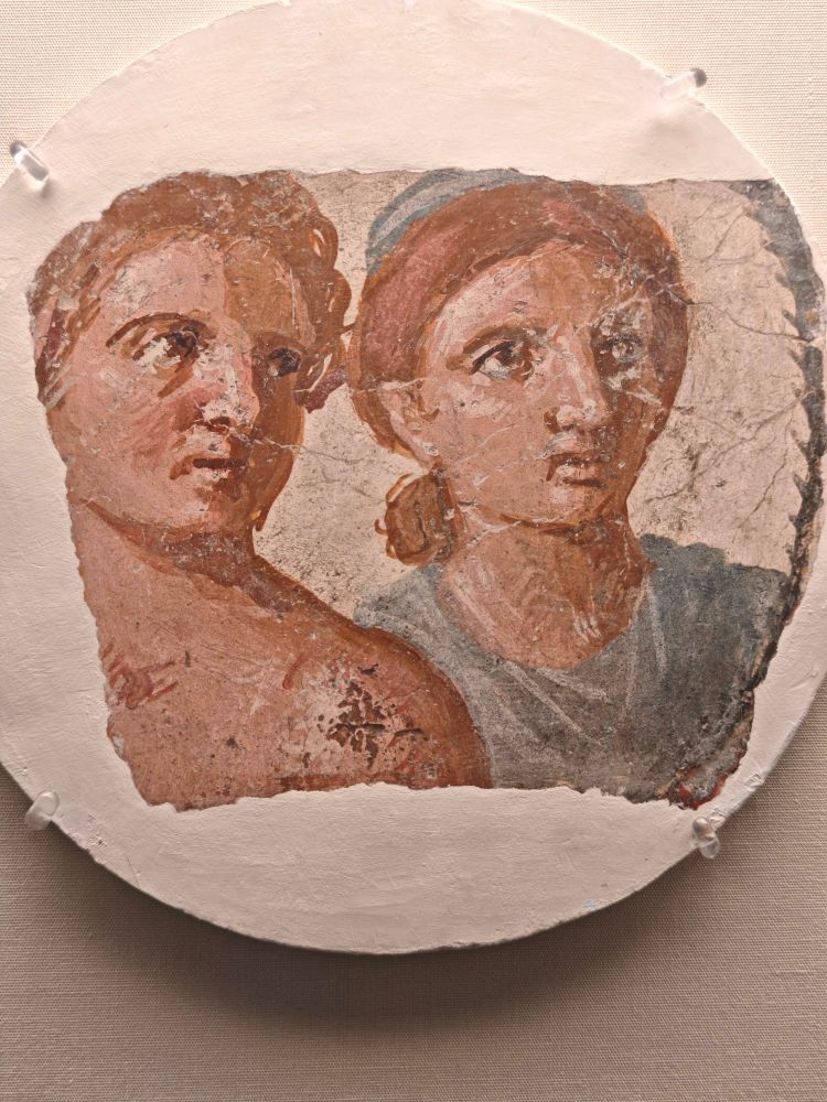 FRESCO ROUNDEL FROM POMPEII, C. 20 BCE - 20 CE. BRITISH MUSEUM

This is part of a genre of wall paintings in Pompeii and Herculaneum, and across the Roman world, images in roundels (part of the leafy frame survives at right) that resemble portrait busts. Sometimes they may depict actual people, perhaps the residents of the house, and sometimes they appear idealised. In this case it's hard to tell. We see here a couple, male and female, looking upward and to our right, in 3/4 view. The male bust at left may be represented as a philosopher with his torso bare, but his face is remarkably individual, his brown hair tousled, his expression verging on apprehension. Next to him, the woman, presumably his wife, has an Augustan-era hairstyle and traces of what might almost be a little hat in blue grey, but is likely a headscarf. She seems idealised. A stop-gap placeholder for a future wife? 