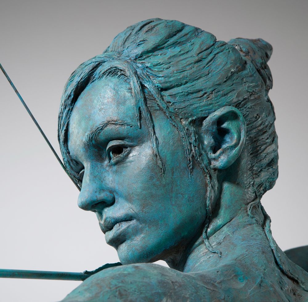 A close up look of the archer’s face from over her shoulder, from the sculpture ’Aiming for Peace’. A white girl in her early 20’s, European features. She looks quietly determined. Her hair tied up in a bun, with some hair on her forehead, and a strand of hair falling passed her eyebrow. You see a tip of her shoulder and there’s a bit of the bow’s string and a bit of the arrow in the picture. She is realistically modelled with as much detail as I could put in to her eyes and lips. Not sure it worked but I wanted the detail to get less described as you got further away from her face. So her ear, is not as detailed as her lips. And not in this photo but by the time you get to the hand holding the bow , it’s more a suggestion. The Blue/Green patina is painted with car spray paints. 