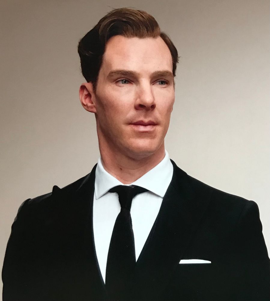 Hard to tell but a wax figure of Benedict Cumberbatch. Very realistic. Bust shot. Black suit with white hanky like a straight line, white shirt black tie. His hair is short back and sides combed over at the front. There’s an auburn tinge to his hair. Background plane, a studio shot. His face looks over the viewer to Benedict’s left. Eyes slightly more to his left. 
