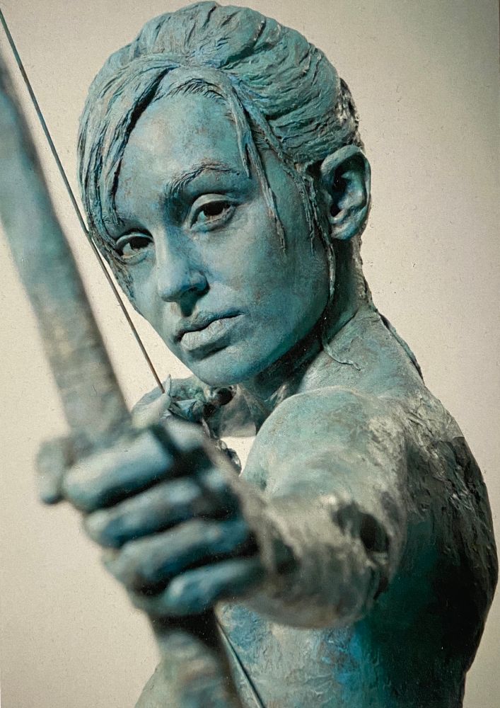 Bust shot of blueish green life size sculpture called ‘Aiming for Peace’. A female archer. Caucasian. Pretty? I think so! Her hair is tied up with some falling on one side of her forehead, and a tiny bit on the other side. She appears to look straight into the camera. Blurred in the foreground, is her fist holding a bow with the arrow pointing directly at us! The bow string running between the bow and the side of her face. This is not clear from the photo, you’d need to know the sculpture. 
The concept of the sculpture is that world peace will require the passion and tireless energy of youth, female energy in full force, and a laser focus like that of an archer. I believe it’s not only possible, but achievable! Merry Christmas, a happy new year and peace on Earth. 