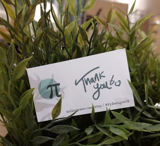 A pydesigns business card that says thank you on it. It's in a bush 