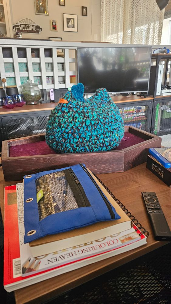 Extra large crochet chicken orb in teal and black sits facing screen left. Chenille fluffy yarn forms the crest on top, and she is otherwise large and proud. 