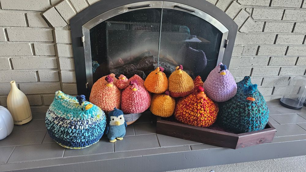 Ten crochet chickens of various sizes and colors sit in front of a fireplace with a single, small penguin nearby. From left to right, a blue white green mixed chicken, a peach orange chicken, a terracotta pink chicken, three orange chickens the colors of various chicken nuggets, an orange and red mixed large chicken, a lilac chicken, and a blue and black chicken. 
The penguin in front is cadet blue with a cream face. 