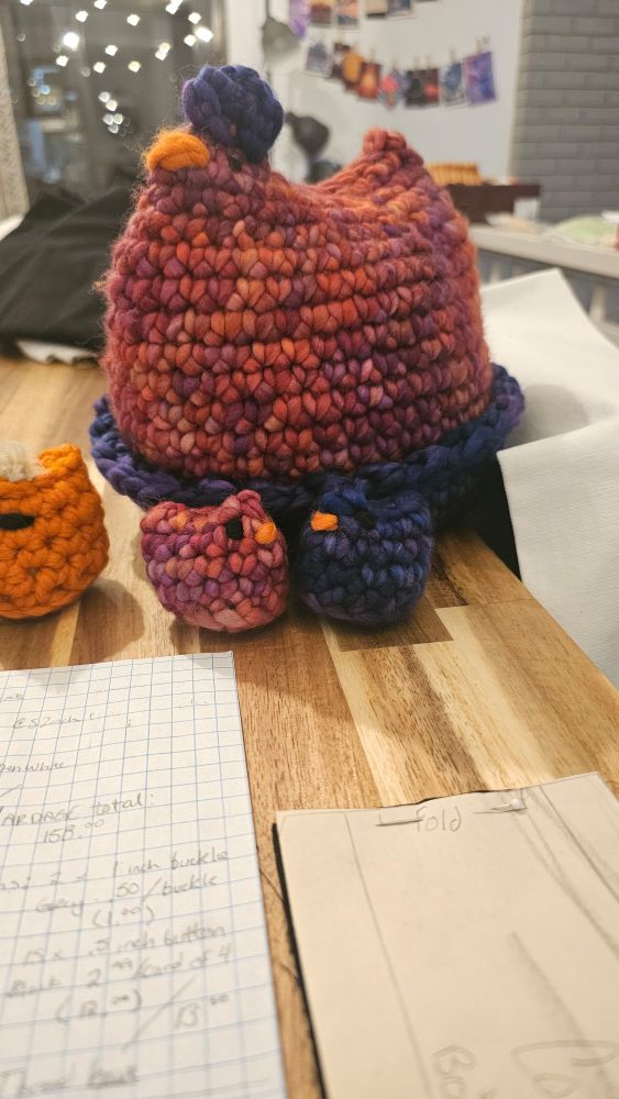 The original finished crochet chicken made of varigated pink/purple/orange yarn. She sits in a nest made of the same kind of yarn but in blue/purple. There are two mini chickens in her yarn colors, and one in a rougher orange yarn sitting around her. She is about ten inches in diameter
