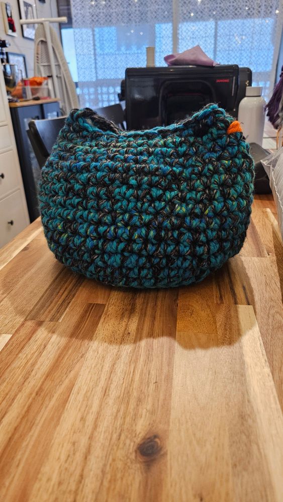 A crochet chicken - orb shaped, low detail- sitting on a kitchen table. The yarn is charcoal and teal, and it has an orange beak. It is a little bigger than a basketball or about a foot in diameter. 