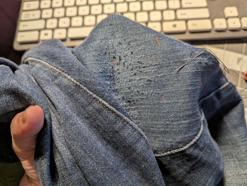 The worn jeans, right side out, focusing on the worn area. The orange thread from the basting stitches is visible around where the patch is. A needle threaded with heavy dark blue thread is stuck through the denim near the edge of the patch. The sewist's left hand is just visible on the left side of the picture -- most of the hand is inside the jeans, supporting the area being sewn.