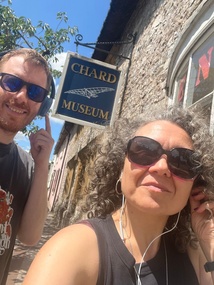 A colour photo of a white man and woman both wearing headphones standing in the Sun outside a pale stone building with a blue sign saying in yellow letters chard museum. 
On the left the man is tall with short ginger hair and a neat moustache wearing sunglasses blue over ear headphones and a dark green T-shirt. On the right a small woman with dark curly shoulder length hair and sunglasses wears a black vest top and has white in ear headphones with cables hanging down. Both are smiling.