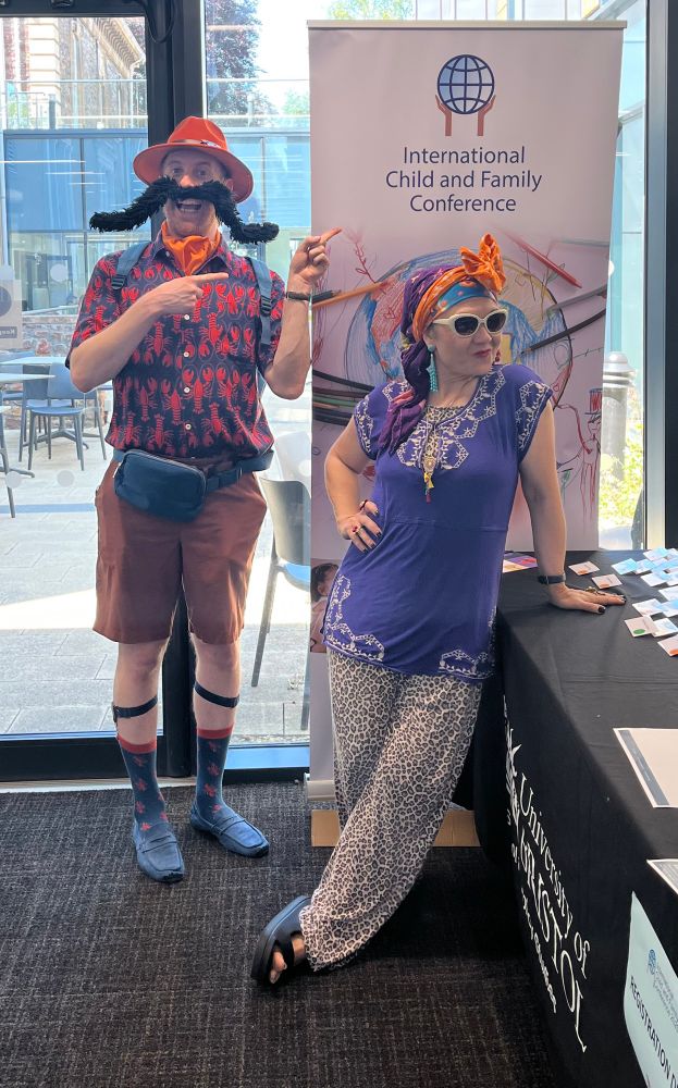 A colour photo of two people standing in front of a long vertical banner. On the left a tall white man in his 30s wearing an orange Fedora hat a large fake bushy black handlebar moustache a short sleeved navy shirt with red lobster print and orange shorts. He is smiling and pointing excitedly at the banner. The banner is white and says in blue international child and family conference. In front of the banner leaning slightly on a table is a small white woman in her 40s. She is wearing an orange head scarf tied in a bow on top of her head, white plastic framed sunglasses and a short sleeved purple tunic with white embroidery on the front over a leopard print jumpsuit.