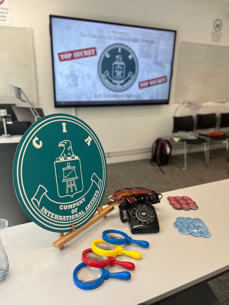 A colour photograph of a seminar room in a university. In the foreground on a white table resting on a table top easel is a teal circular logo with white writing saying CIA company of international artists. Next to the logo on the right black old-fashioned telephone with a lobster attached to the handset. Also on the table several plastic magnifying glasses in red yellow and blue. Slightly out of focus on the wall behind a PowerPoint presentation with the same circular teal logo in the centre and the words top-secret in red can just be seen.