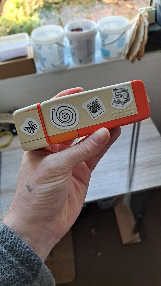 my hand holding a small beige and orange 110 camera, now adorned with life is strange icon stickers - a butterfly, the iconic spiral icon, a polaroid photograph and a polaroid camera.