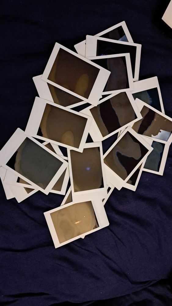 dozens of instant photos lie on a blue surface. they're all mostly black due to underexposure.