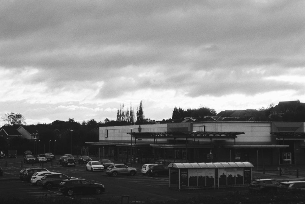 a grainy black and white photograph of a large supermarket with a car park mostly full of cars. the sky is cloudy and dark.