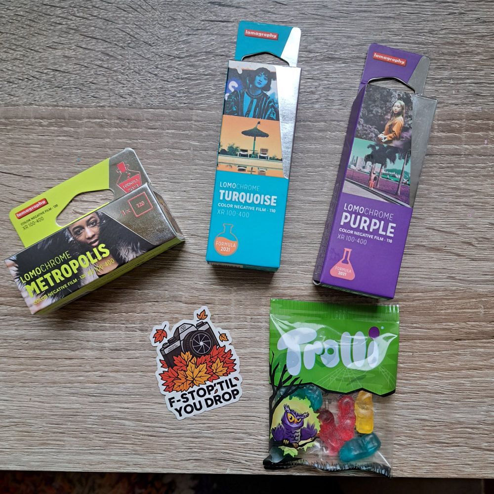 a table on which lies 3 films - lomochrome metropolis in 120, and lomochromes purple and turquoise in 110 - as well as a sticker depicting a camera surrounded by autumn leaves with the slogan "F-STOP 'TIL YOU DROP" and a bag of gummy worms.