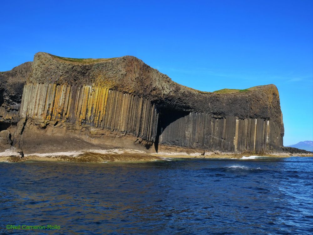 The island of Staffa viewed from the sea, in the centre the entrance to McKinnon's Cave (one of several caves on the island, including Fingal's Cave) casts a shadow on the Basalt columns. The sea is dark blue beneath a bright blue sky.

The spectacular columns of basalt run across the whole of the island in this view. They are sandwiched between pyroclastic rocks at the bottom and a second volcanic flow at the top (this cooled more rapidly than the middle layer so failed to form columns)