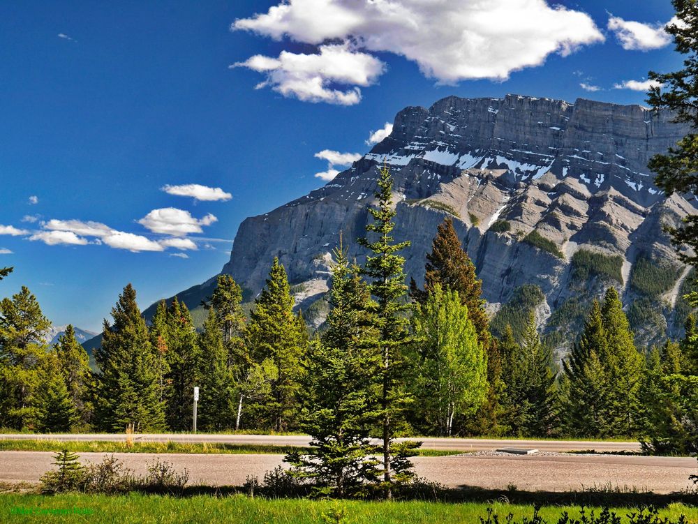 Looking South from the campsite, thick conifer forest grows on the edge of the Bow River Valley, on the other side of the valley the near vertical face of Mount Rundle rises majestically towards the sky. The bedding of the sedimentary rocks can be clearly seen pitched at an angle of about 30Deg (rising from the left), snow has accumulated on the ledges near the top. All beneath a bright blue sky dotted with fluffy white clouds.