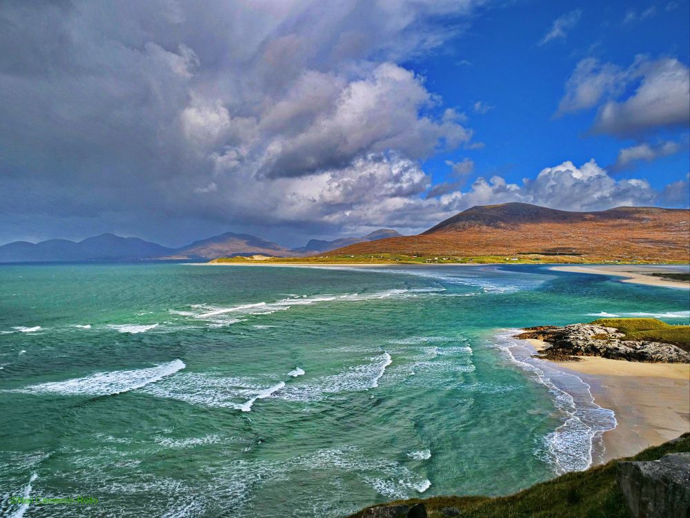 Taken from a rise above Seilebost Beach the turquoise waters of the Atlantic send waves to crash upon the beach. In the middle distance a wides shallow bay stretches towards the start of Luskentyre Beach which is in the centre of the image. In the distance the rugged hills of Harris dominate the horizon. Above, stormy looking clouds on the left herald the arrival of a rain squall, to the right blue sky with fluffy clouds allows fpr patches of sunlight to illuminate the scene.