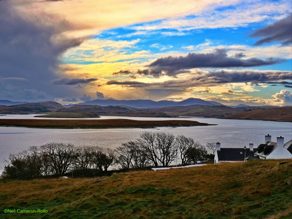 Looking South across the waters of a sea loch towards the hills of North Harris on the horizon. The loch is battleship grey and the landscape is muted shades of brown and green.
In the sky to the left angry clouds from a squall that has just passed fill the view. The squall leaves behind a turbulent sky with patches of blue, the clouds are tinged with orange from the low Autumn Sun