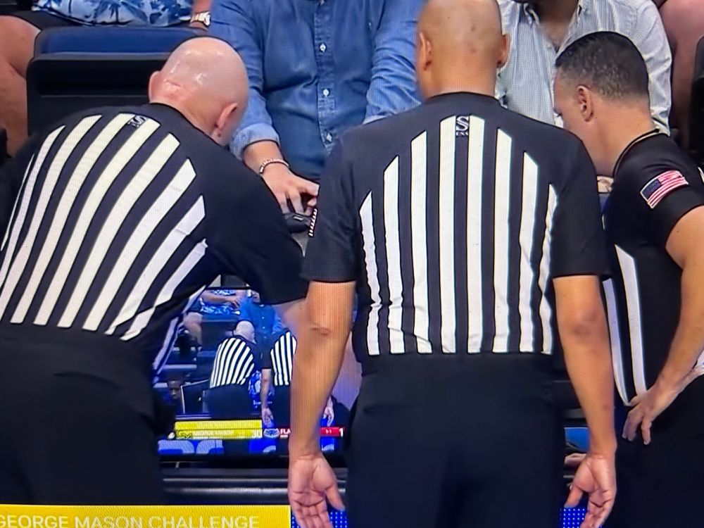 3 college basketball refs stand at the scorers table looking at a monitor. The monitor is showing the game broadcast which is showing the refs.  