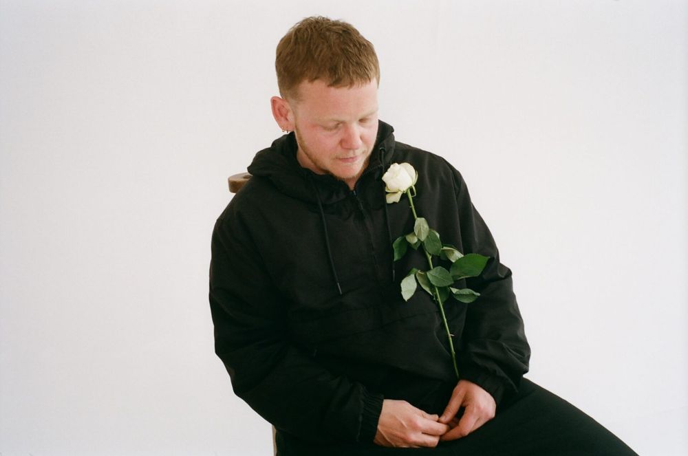 Photograph of Kae Tempest, dressed in black and holding a single white rose. 