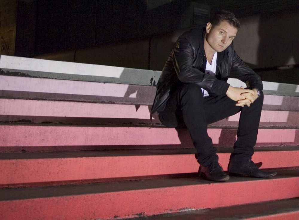 Douglas McCarthy of Nitzer Ebb, wearing all black apart from a white shirt, sat on a set of rising steps. 