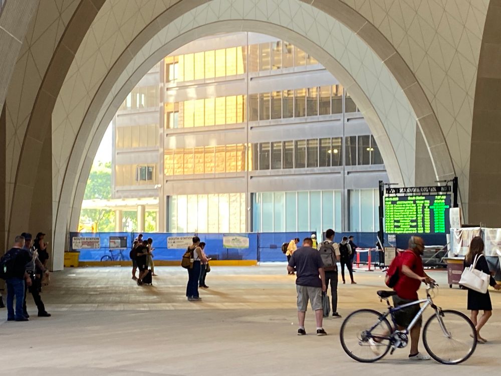 A view of the area between South Station building and the train platforms. The arches of the new South Station Tower frame people and a background building 