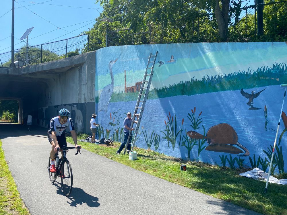 A person riding a bicycle passes a team painting a mural